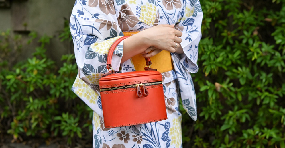 橘紅色皮革圓筒型手提包，配有雙拉鍊開合及提把，整體風格簡潔。