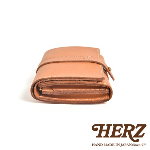 這是一個由日本品牌 HERZ 職人手工製作的皮革皮夾，以側視角呈現其厚實且富有層次的細節。皮夾選用質感溫潤的淺棕色天然皮革，表面帶有細緻的皮革紋理，邊緣處呈現明顯的堆疊層次。皮夾中間有一條帶有縫線細節的皮革壓帶，並配有金屬釦具固定，整體呈現簡約且經典的手作質感，右下角印有 HERZ 品牌標誌與相關手工藝標語。