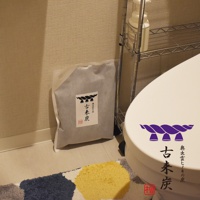 灰色包裝袋裝有「古來炭」字樣和繩結圖案，旁邊為白色馬桶，上方置物架上有白色瓶裝物。