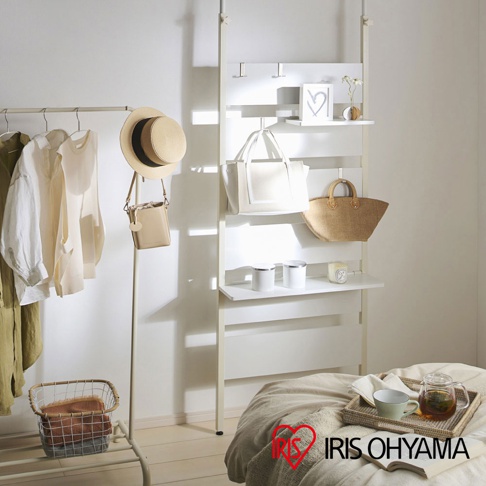 頂天立地收納架，白色木質層架上掛有白色托特包、草編包，另有裝飾畫框、花瓶、罐子和香氛蠟燭。左側掛衣架上有多件淺色衣物和一頂草帽。