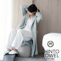 一位女性穿著淺藍色蓬鬆浴袍，內搭白色上衣與白色長褲，坐在灰白色圓形坐墊上，雙手正在整理頭髮。