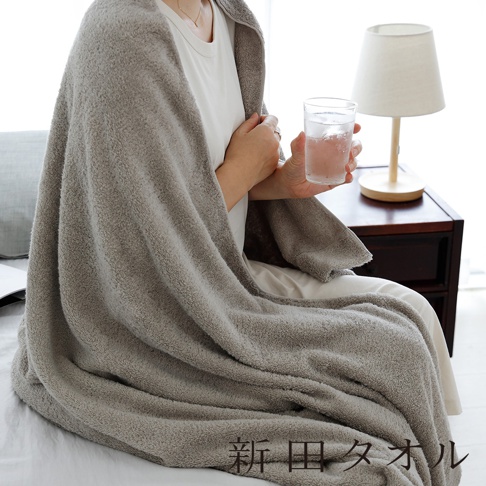 一位女性坐在床上，披著一件觸感柔軟、質地蓬鬆的灰褐色毛巾單人被，手中拿著一杯水，右下方印有新田タオル的字樣，整體呈現舒適放鬆的居家氛圍。