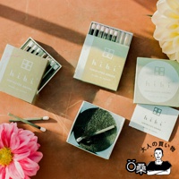 香氛火柴禮盒，包含多個淺綠色紙盒，上面印有「hibi」品牌標誌及「10MINUTES AROMA MADE IN JAPAN」字樣。盒內裝有黑色木桿、白色火柴頭的香氛火柴，部分火柴已點燃。另有兩個綠色圓形貼紙，同樣印有品牌標誌。產品背景為橘棕色桌面，左側有粉紅色大朵花卉，右側有淡黃色花朵。