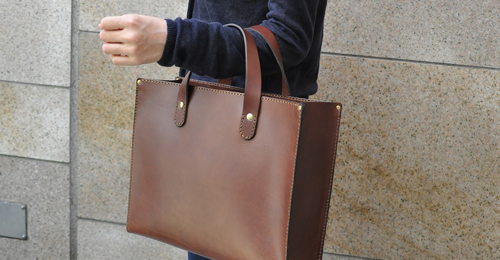 職人手作皮革直式托特包，咖啡棕色，光滑皮革材質，簡潔設計，配有兩個手提把，包身側面與頂部邊緣有縫線裝飾，並有金屬卯釘固定。