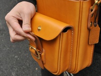 職人手作皮革皮扣直式單肩包，採用天然鞣製皮革，呈現溫潤的焦糖色。包身結構清晰，縫線紮實，配有金屬皮扣及可調節的皮革背帶，展現簡約實用的設計風格。