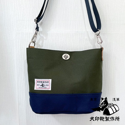 犬印純綿帆布雙色迷你側背包，主體為橄欖綠，底部為海軍藍。包包正面有品牌標籤，配有銀色金屬旋轉鎖扣和深藍色帆布肩帶，肩帶可調節。