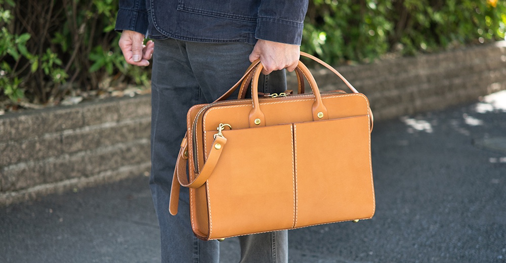 棕褐色皮革雙口袋兩用公事包，有頂部提把及可拆卸肩帶，黃銅色拉鍊及金屬配件，縫線清晰。