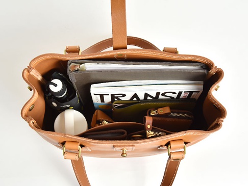 職人手作皮革原子釦托特包，淺棕色天然皮革製成，包內裝有文件夾、雜誌、錢包、摺疊傘和白色圓形物品，展現包款的收納空間與質感。