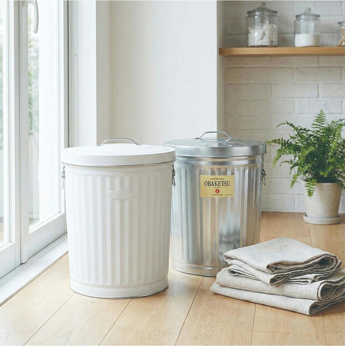 OBAKETSU 品牌白色和鍍鋅鋼垃圾桶，帶有把手和蓋子，擺放在廚房檯面上。
