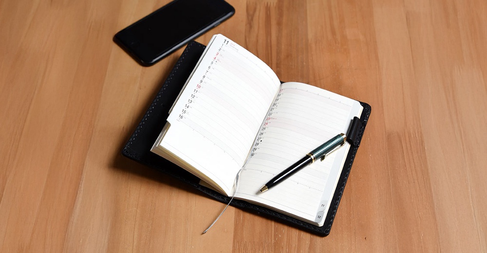 黑色皮革筆記本書套，內含一本攤開的行事曆，筆記本中央夾著一支綠色與金色的鋼筆，上方另有一支黑色手機，皆放置於木紋桌面上。