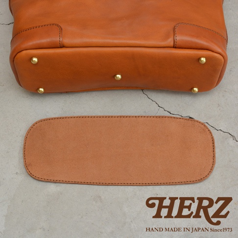 HERZ 品牌標誌，下方文字為 HAND MADE IN JAPAN Since1973，旁邊是棕色皮革托特包的底部細節，以及一個與之搭配的皮革襯墊。