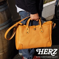 職人手作皮革大容量托特包 T-146，採用天然皮革製成，顏色為溫暖的淺棕色，包身結構紮實，配有雙提把與可拆卸肩背帶，表面有精緻縫線與金屬卯釘裝飾。