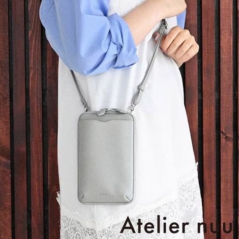 一位女性身穿白色蕾絲邊上衣搭配藍色襯衫，斜背著一款 Atelier nuu 品牌的淺灰色皮質隨身手機包，包款設計簡約並配有細背帶，背景為深褐色直條紋木質牆面。