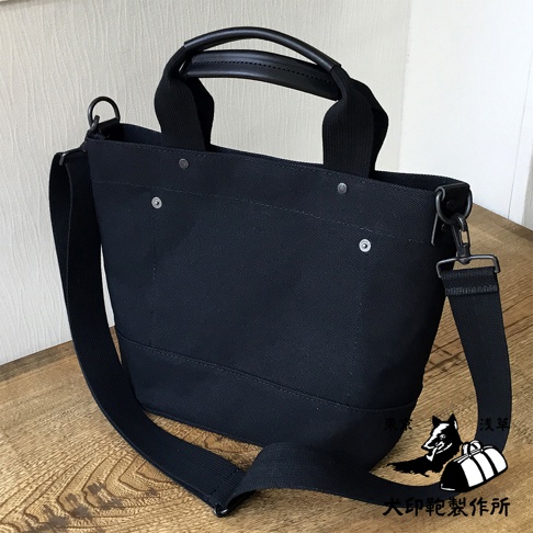 黑色帆布兩用托特包，附有皮革提把與可拆卸的帆布肩背帶，包身綴有金屬鉚釘。