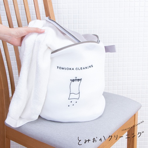 白色網狀浴巾洗衣袋，袋身印有「TOMIOKA CLEANING」字樣及衣物滴水的插畫。袋口有灰色拉鍊與提把，一條白色浴巾部分放入袋中。