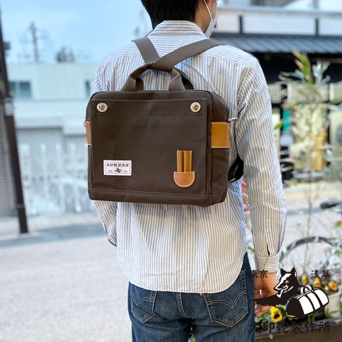 深棕色純棉帆布製成的三用側背包，搭配皮革飾邊，背包帶肩背於身穿藍白條紋襯衫的男子身上。