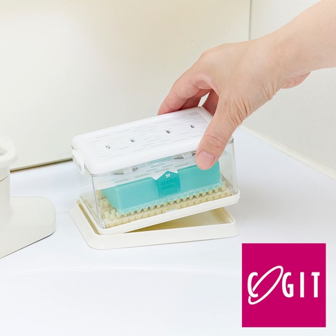 透明塑膠盒內置藍綠色洗衣皂，底部有白色濾網，盒蓋為白色，上方有條狀透氣孔。整個結構置於淺盤上，一隻手正在蓋上蓋子。