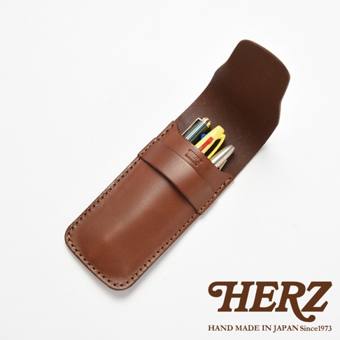 這款日本HERZ職人手作皮革鋼筆收納筆袋呈現棕色皮革質感，採直立式開口設計，筆袋前方印有HERZ品牌標誌，內部插有三支不同顏色的鋼筆，整體風格簡約且充滿職人手作細節，右下角配有HERZ品牌文字與創立年份資訊。