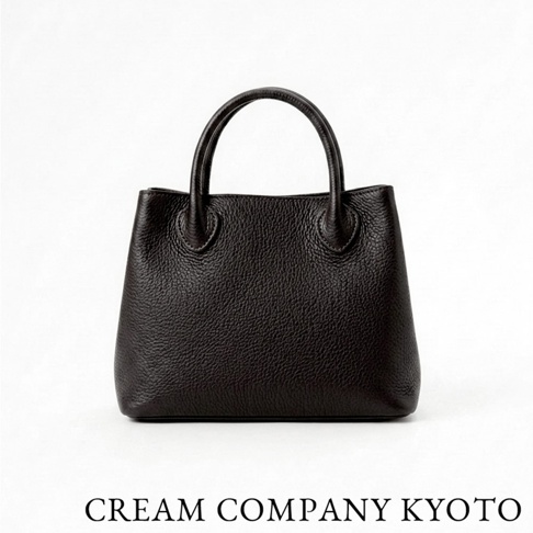 一款由 CREAM COMPANY KYOTO 推出的深棕色皮革手提托特包，包身採用帶有自然紋理的牛皮革製作，外型簡約典雅，正面視角展示其挺立的包型與精緻的圓弧提把設計，下方印有品牌名稱。