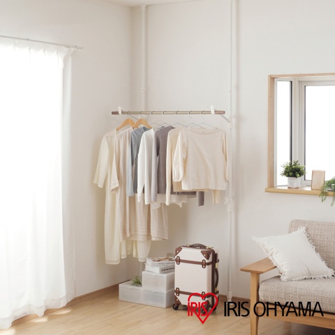 白色頂天立地曬衣架，頂部有木紋飾條，掛有多件淺色系的衣物，下方有白色行李箱及書本堆疊。