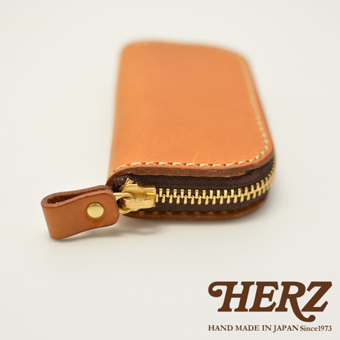職人手作皮革橢圓形拉鍊鑰匙包，淺棕色皮革，附金色拉鍊和皮革拉環。下方有HERZ品牌標誌及 HAND MADE IN JAPAN Since1973 字樣。