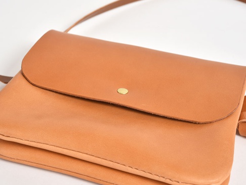 職人手作皮革直式隨行袋，採用光滑的淺棕色皮革製成，帶有翻蓋設計和一個黃銅色金屬鈕扣。