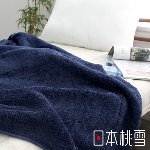 深藍色今治飯店毛巾被，材質柔軟，觸感舒適，覆蓋在床上，一旁有白色枕頭和雜誌，背景有綠色植物裝飾。