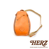 一皮革製斜背包，呈水滴狀，表面光滑，帶有拉鍊開合，左側有一傾斜拉鍊口袋，前方中央有一個菱形皮革標籤，附有可調節的帆布與皮革混搭肩帶，顏色為橘棕色。