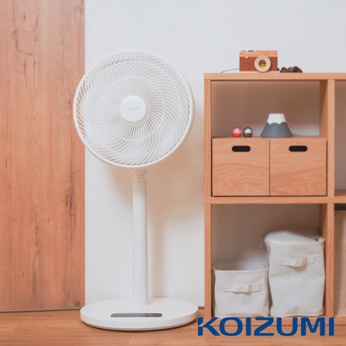 放置在室內木質地板上的日本KOIZUMI純淨白DC變頻遙控電風扇，風扇主體為極簡圓形設計並搭配纖細支架，右側放置一座淺木色開放式層架，架上陳列著木製相機模型與裝飾擺件，整體空間呈現溫馨自然的居家風格。