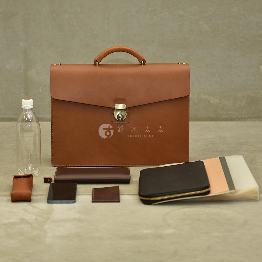 職人手作的棕色皮革公事包，配有金色金屬鎖扣和提把，旁有水瓶、卡夾、手機、長夾和一個拉鍊收納包。