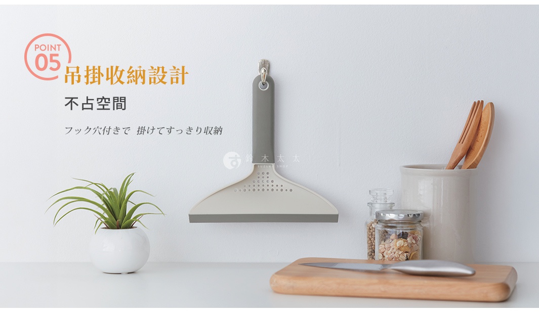 一個奶油色與灰色調的清潔刮鏟掛在白色牆面上，手柄頂端設計有孔洞方便吊掛收納，前方桌面上擺放著小盆栽、裝有穀物的玻璃罐以及木製砧板與刀具，整體呈現簡約的生活家居風格。