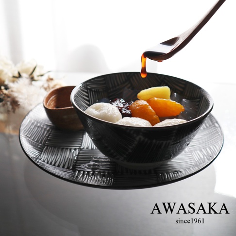 黑色和風紋飾的陶瓷碗盛著日式甜點，碗內有白色湯圓、橘色糖漬水果塊、黃色水果塊與黑色寒天凍，上方有一個木勺正滴落糖漿，碗置於同款紋飾的淺盤上。