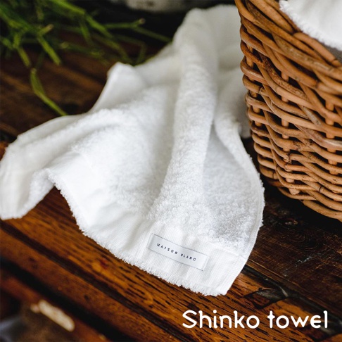 MAISON BLANC 標籤的白色柔軟匹馬棉浴巾，有著細緻的毛圈織紋，搭配藤編籃和綠色植物，陳設在木質桌面上。