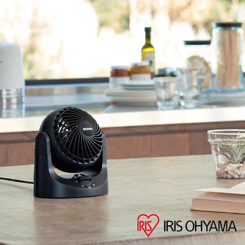 黑色桌上型空氣循環扇，扇葉網罩有螺旋紋路，底座有旋鈕開關，放置於木紋檯面上，背景為模糊的廚房窗戶與餐具。
