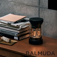 The BALMUDA wireless speaker is a cylindrical device with a clear glass body, revealing two illuminated vacuum tubes inside. It features a black grille on top and a black base.