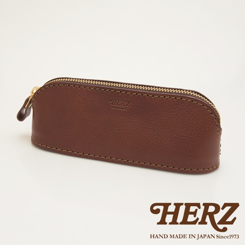 職人手作皮革三角弧形收納筆袋，採用棕色皮革製成，設有金色拉鍊和品牌壓印。