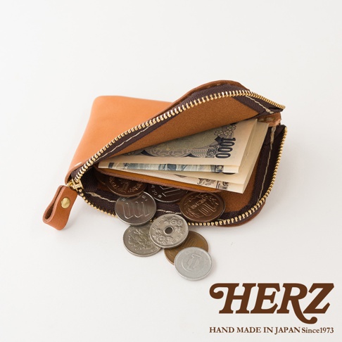 一個呈現開啟狀態的棕色職人手作皮革迷你零錢包，內部收納了幾張一千日圓紙鈔及數枚日本硬幣，拉鍊細節精緻，右下角印有日本HERZ品牌標誌，整體風格簡約且極具質感。