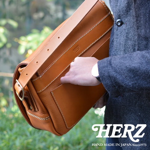 職人手作復古翻蓋郵差包，採用棕色皮革材質，配有可調節的肩帶和金屬扣環，包身車縫線明顯，展現精緻工藝。