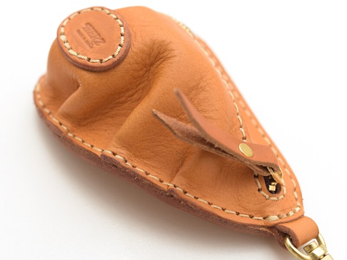 職人手作皮革餃子造型零錢包，採用天然棕色皮革，以白色縫線手工縫製，附有黃銅色拉鍊和掛環，頂部圓形皮革上印有「HERZ」品牌標誌。