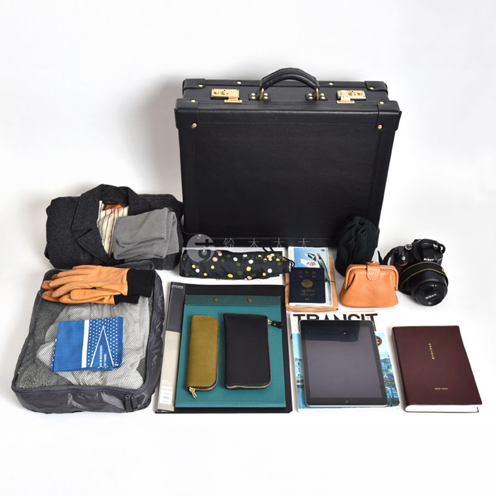 黑色皮革手提行李箱，配有金色鎖扣和提把，周圍散落著衣物、皮手套、摺疊傘、護照、錢包、數位相機、平板電腦和筆記本。