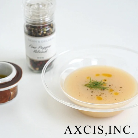 清透玻璃深型點心碗，盛有淺黃色湯品，上方點綴綠色香草和黑胡椒粒，碗旁為一瓶綜合胡椒粒研磨器及一個深褐色陶瓷小碟。