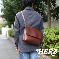 一位男性身穿深藍色外套，斜背一個棕色皮革經典郵差包。包款設計簡約，具有一個較大的翻蓋，皮革質感光滑。