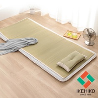 淺棕色藺草編織的單人涼蓆，鋪設在木質地板上，蓆面上有灰色毛巾和一個淺綠色枕頭，角落有品牌標誌。