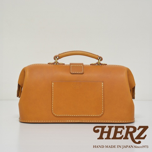 職人手作皮革長型迷你醫生包，採用天然皮革材質，呈現溫潤的焦糖棕色。包款設計經典，擁有頂部提把、金屬釦環與前置方形皮夾層，整體線條簡潔，展現手工的細緻與質感。