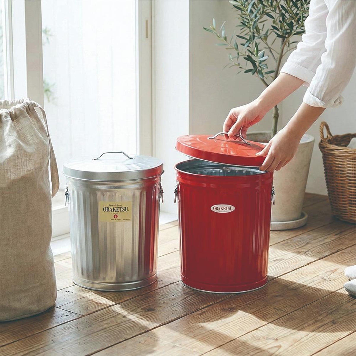 一個紅色 OBAKETSU 垃圾桶，上方蓋子由人的手拿起，旁邊有一個銀色 OBAKETSU 垃圾桶，還有一個麻布袋。