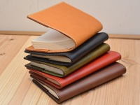 堆疊的職人手作皮革厚革書套，從上至下顏色分別為棕褐色、黑色、墨綠色、紅色、淺棕色。頂部書套露出部分白色紙張，皮革邊緣有縫線裝飾。