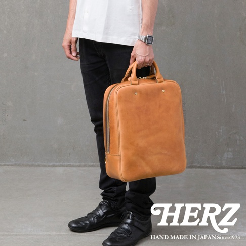 HERZ 品牌標誌，下方為 HAND MADE IN JAPAN Since1973 字樣。一個人手提著一個棕色皮革後背包，他穿著白色襯衫、黑色長褲和黑色休閒鞋。