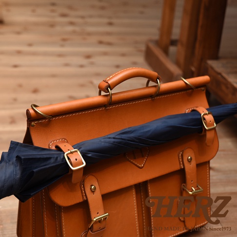 職人手作皮革旅行包 A-102，採用天然皮革，呈現溫潤的棕色調。包款設計簡約，配有可調整的皮質揹帶和金屬釦環，正面有兩個翻蓋口袋，並以精緻的縫線裝飾。包包上固定著一支深藍色的摺疊傘。