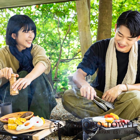 一男一女在戶外野餐，女子身穿卡其色上衣搭配深藍色圍巾，男子身穿黑色上衣搭配米白色圍巾，兩人面前有烤肉架、食材及木盤。