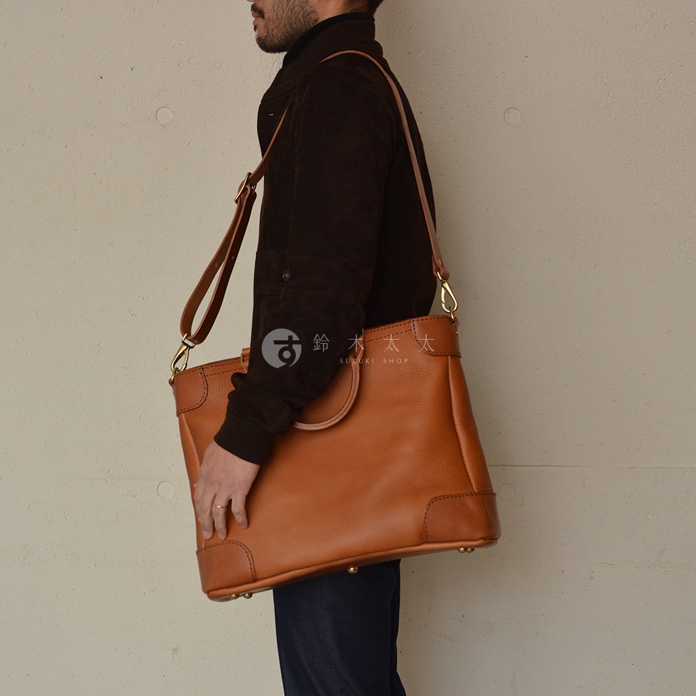 棕色皮革製成的兩用商務托特包，包包有提把和可拆卸的長背帶，搭配金色五金配件，包包底部有四個金屬腳釘。